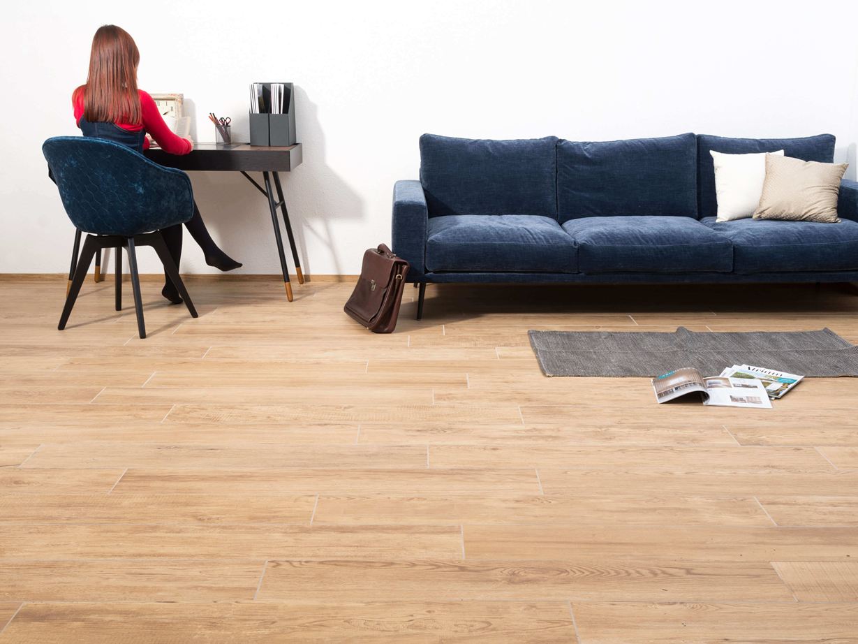 Femme assise à table à côté d'un canapé dans un salon avec des carreaux de grès cérame dans un aspect bois comme revêtement de sol.