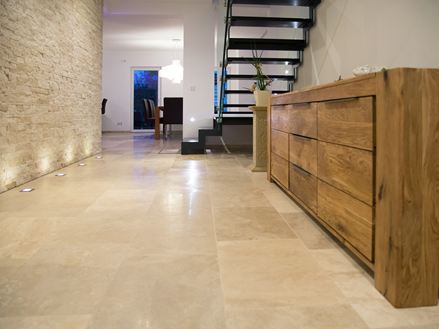 Un sol en travertin sur la droite un commode en bois en arrière plan un mur sur la gauche et des escaliers noirs à droite au fond la cuisine