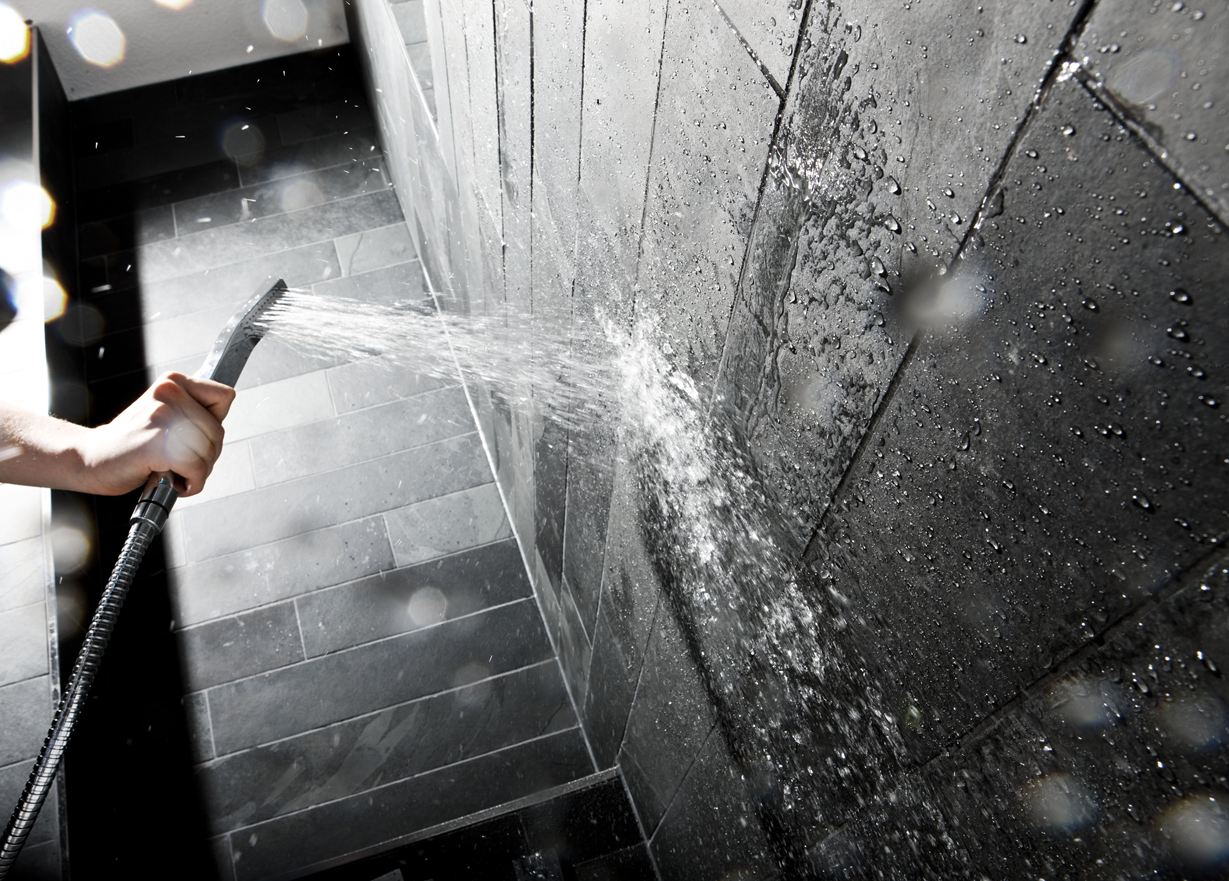 Hand hält eine Duschbrause gegen eine Wand mit dunklen Steinfliesen