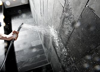 Hand hält eine Duschbrause gegen eine Wand mit dunklen Steinfliesen